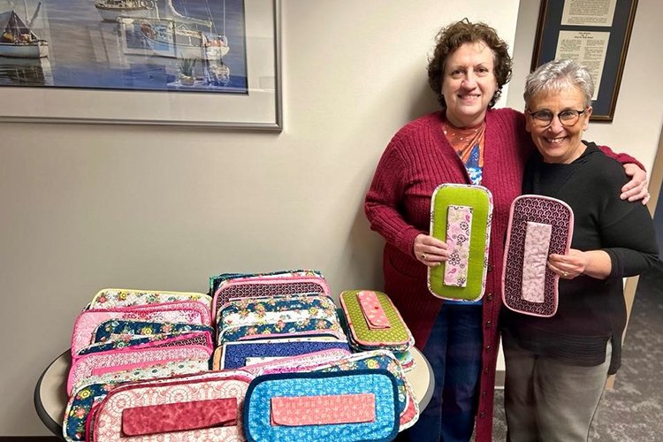 Image of two women standing by a table of hand quilted bosom buddies. 