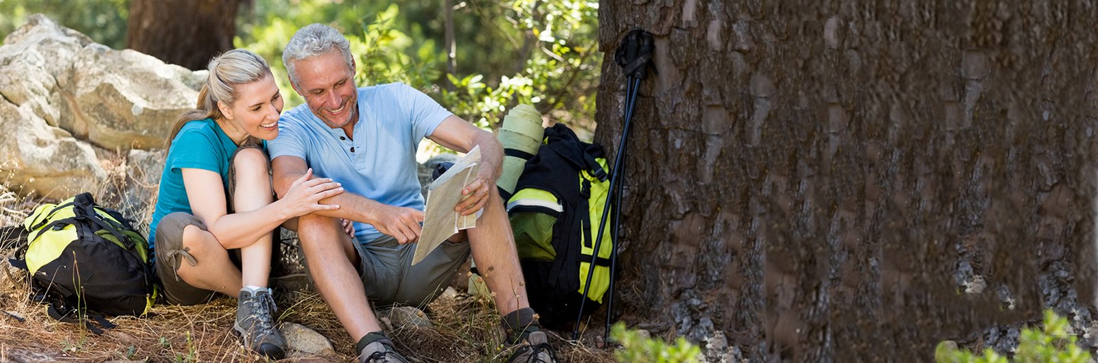 Photo of a man and woman sitting by a treat in hiking clothes looking at a map.