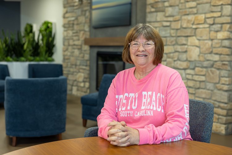 Picture of Patsey Deveney sitting at a table with fireplace behind her, smiling at the camera.